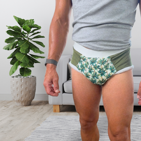 A man wearing patterned green and white adult cloth diaper with a white waistband, standing in a living room next to a potted plant.