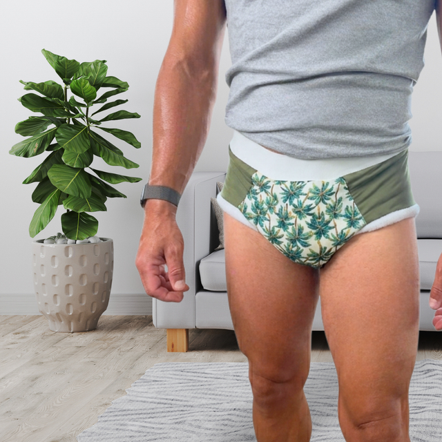 A man wearing patterned green and white adult cloth diaper with a white waistband, standing in a living room next to a potted plant.