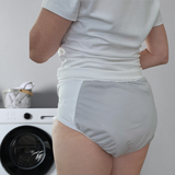 A women wearing light gray incontinence briefs or adult diaper, standing in front of a white washing machine in a clean indoor setting.